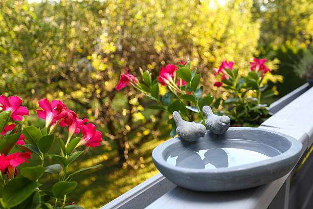 Small grey ceramic bird bath with bird figurines sits on a balcony, surrounded by pink flowers.